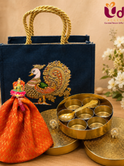 Premium Varalakshmi Vratham return gift hamper featuring a peacock printed jute bag, brass masala box with seven compartments and spoon, brocade potli bag, and lotus haldi kumkum.