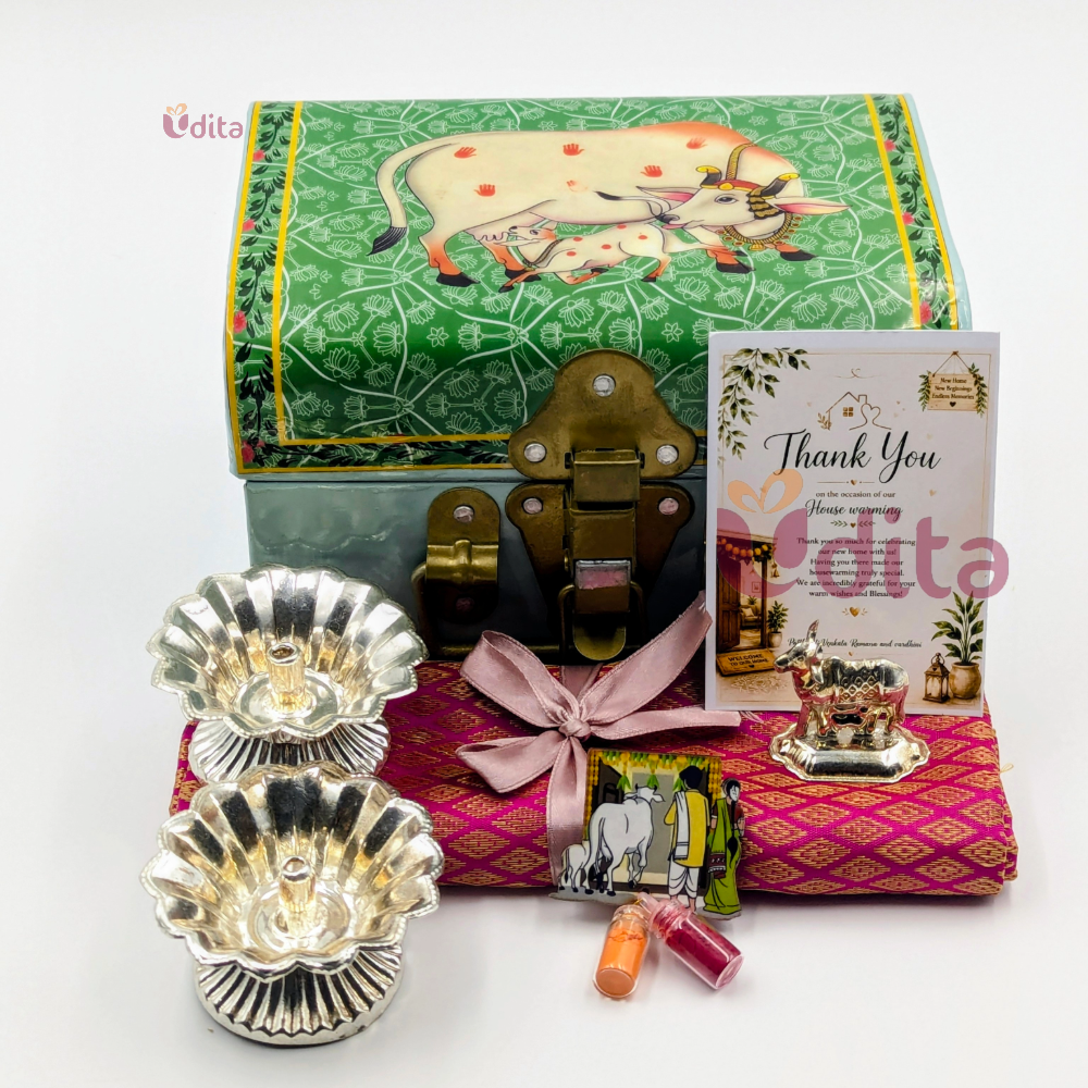 Pichwai trunk box housewarming hamper with German Silver diyas, Kamadhenu idol, brocade blouse piece, and thank you card.